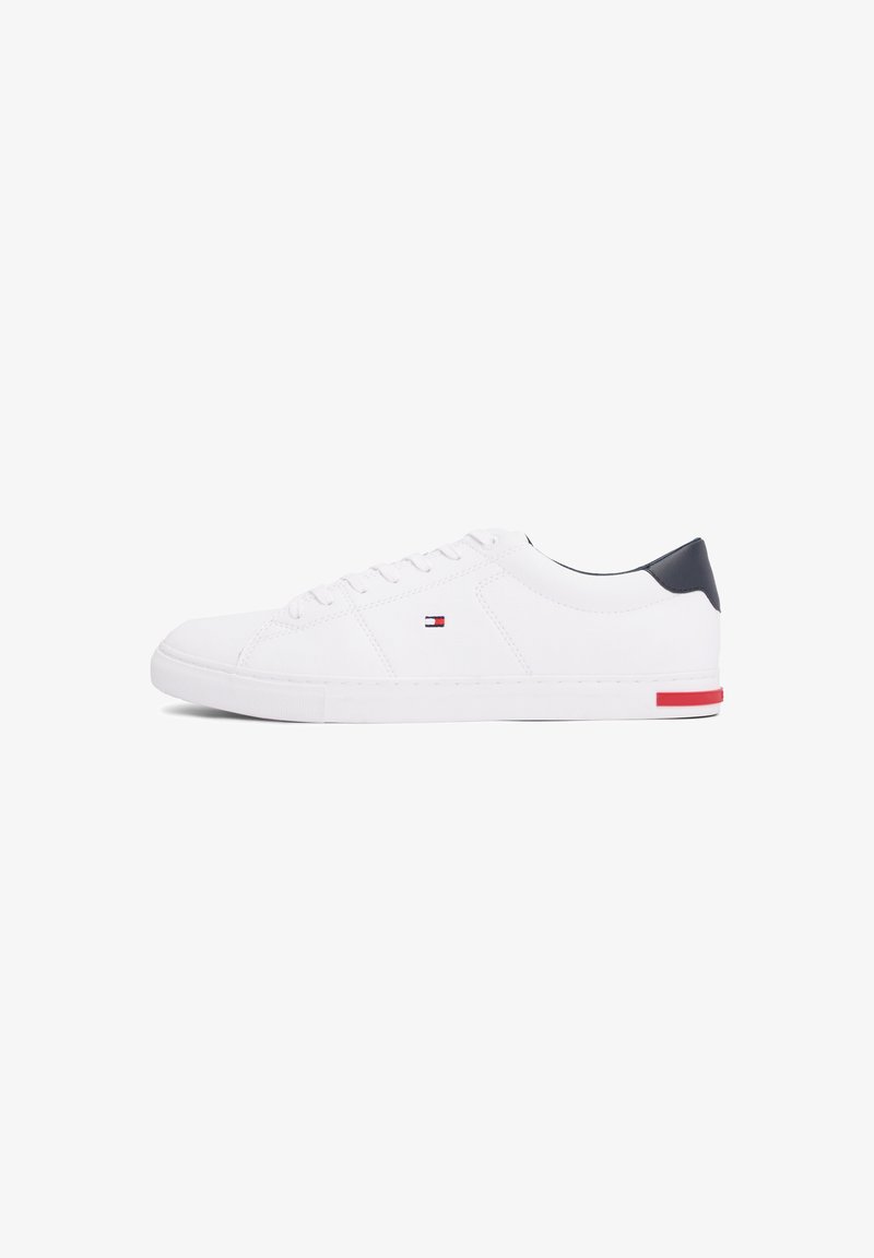 Baskets basses blanches avec languette de talon bleu marine, petit logo rouge et bleu sur le côté, lacets blancs et une bande rouge sur la semelle près du talon.