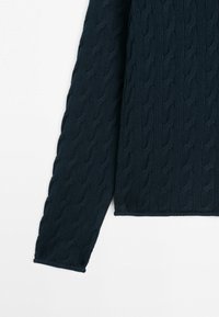 Ærme af en marineblå strikket sweater med et kabelmønster på en hvid baggrund, der viser en del af overkroppen og armen.