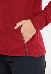 Röd fleece-pullover med en dragkedjeficka. Tyget är mjukt, strukturerat, har en figursydd design och en svart accent på dragkedjan.