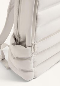 Világos bézs hátizsák párnázott textúrával, dupla cipzáras rekeszekkel és állítható pántokkal a kényelmes és funkcionális használat érdekében.