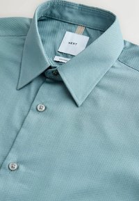 Chemise en coton texturé bleu clair avec col pointu et boutons blancs, pliée avec l'étiquette de marque visible « NEXT ».