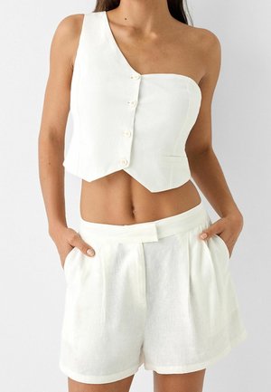 Kobieta ubrana w biały asymetryczny, jedno-ramienny crop top z guzikami, zestawiony z wysokim stanem białych plisowanych szortów, ręce w kieszeniach.