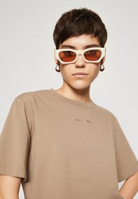 Sonnenbrille mit weißem Rahmen und braunen Gläsern, kombiniert mit einem beigen Baumwoll-T-Shirt mit kleinen, gestickten Texten am Halsausschnitt.