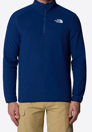 Mężczyzna noszący granatowy polar z długim rękawem na ćwierć zamka z białym logo The North Face na lewej piersi oraz beżowe spodnie typu cargo.