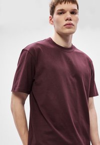 Bordo bombažna majica z okroglim izrezom, kratkimi rokavi in subtilnim vtisnjenim logotipom na prsih. Enostaven dizajn, gladka tekstura.