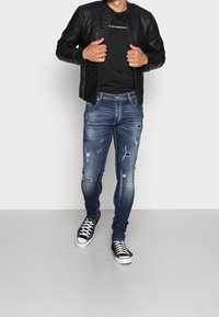 Giacca bomber in pelle nera, t-shirt nera, jeans skinny blu strappati con schizzi di vernice e sneakers in tela bianco e nero.