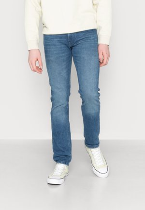 Persoon draagt medium blauwe slim-fit jeans, off-white sweatshirt en off-white lage canvas sneakers, staand op een grijze vloer.