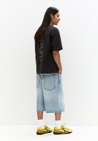 Black short-sleeve T-shirt with white vertical text; paired with light blue denim shorts and yellow sneakers. Smooth fabric, casual fit.