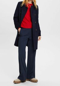 Marineblauer langer Mantel mit goldenen Knöpfen über einem roten Pullover, kombiniert mit dunkelblauen weit geschnittenen Hosen und beigen klobigen Schuhen mit dicker Sohle.