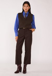 Gilet di lana marrone indossato sopra una camicia blu, abbinato a pantaloni scuri a righe sottili e stivali bordeaux con tacco. Accessori comprendono anelli e cintura.