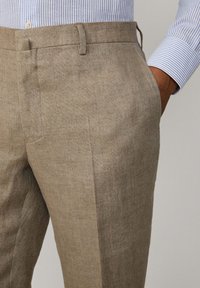 Tan linen trousers with a smooth texture, featuring a straight cut, side pockets, and a flat front design. Paired with a blue striped shirt.