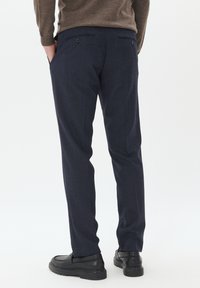 Námornické kalhoty z texturované látky, s zadními kapsami se dvěma knoflíky, nošeny s černými botami, viděny ze zadu.