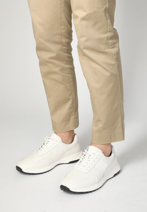 Weiße Leder-Sneaker mit runder Zehenpartie, flacher schwarzer Sohle und klassischem Schnürdesign, kombiniert mit beigen, taillierten Hosen.