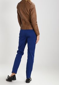 Bruine leren jas met een aansluitend ontwerp, gecombineerd met blauwe broek met een rechte snit. Zwarte schoenen met een strikdetail maken de outfit compleet.