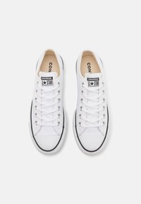Weiße Canvas-Sneaker mit schwarzen Gummisohlen, ausgestattet mit weißen Schnürsenkeln und einem schwarzen Logo-Patch auf der Zunge. Einfaches, niedrig geschnittenes Design.