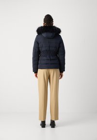 Doudoune marine avec capuche en fausse fourrure, taille froncée et texture matelassée. Associée à un pantalon beige à jambes larges et des bottines noires.