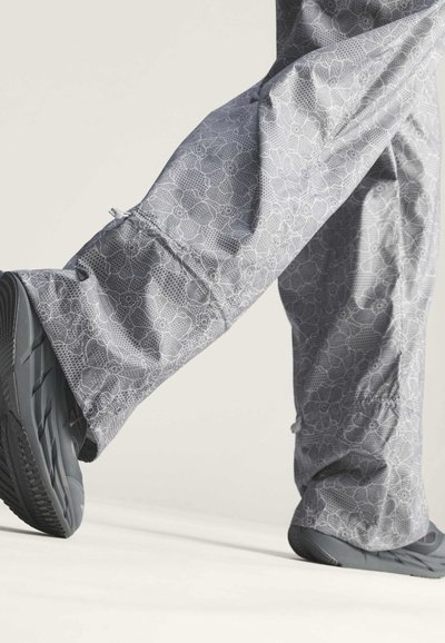 Jambes portant un pantalon gris à motif floral et des chaussures noires, un pied levé comme si on marchait sur une surface claire.