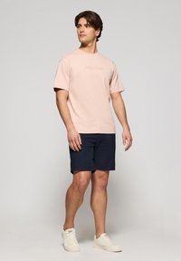 Camiseta de algodón rosa claro con el texto "Polo Club", combinada con pantalones cortos marinos y zapatillas blancas. El modelo está de pie con los brazos relajados a los lados.