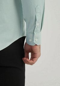 Chemise texturée vert clair avec des poignets à boutons, des coutures détaillées et un ourlet incurvé, associée à un pantalon sombre, montrant une partie d'une main.