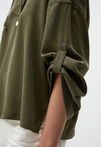 Pullover oversized verde olivo con maniche arrotolate. Presenta una texture liscia, spacchi laterali e dettagli in metallo argentato.