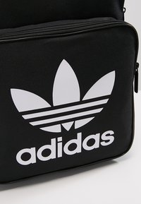 Black backpack with a smooth texture; features a prominent white Adidas logo and three horizontal stripes. Zipper closure at the top.
