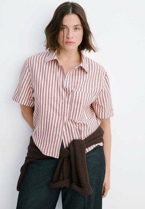 Jeune femme aux cheveux courts bruns portant un t-shirt à manches courtes rayé rouge et blanc et un pantalon foncé avec un pull marron noué autour de la taille.