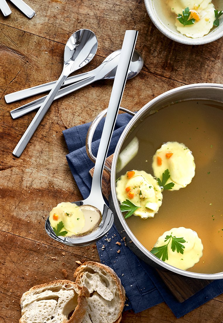 Łyżka ze stali nierdzewnej nabierająca zupę z pierożkami, położona obok niebieskiego materiału, z plastrami chleba na drewnianej powierzchni.