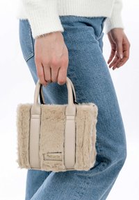 Beige nepbont handtas met twee bijpassende leren handvatten, rechthoekige vorm en een gouden merketiket aan de voorkant, vastgehouden door een persoon.