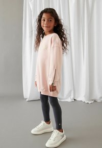 Růžová oversized mikina s hvězdným detailem, se zipovými manžetami, v kombinaci s tmavými žebrovanými legínami a bílými teniskami se stříbrnými akcenty.