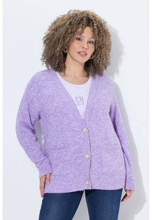 Frau mit lockigem Haar trägt eine helllila Strickjacke mit Knöpfen über einem weißen Shirt mit Aufdruck und eine blaue Jeans, stehend vor einem schlichten Hintergrund.