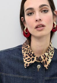 Veste en jean avec un col imprimé léopard, dotée d'une fermeture à boutons sur le devant et de grandes poches. Portée avec des boucles d'oreilles circulaires rouges.