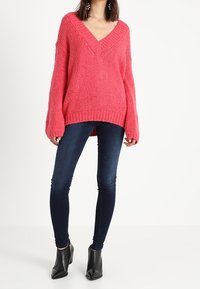 Suéter de punto rosa con escote en V y mangas oversized, combinado con jeans skinny azul oscuro y botines negros de tacón, sobre un fondo blanco.