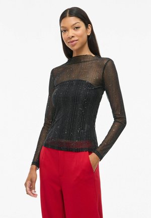 Zwarte doorschijnende top met lange mouwen, horizontale strepen en subtiele sparkle. Hoge hals en aangesloten vorm, gecombineerd met rode wijde broek.