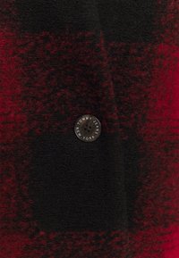 Wełniana tkanina w kratę w kolorach czarnym i czerwonym, z okrągłym czarnym guzikiem z wygrawerowanym napisem "TOMMY JEANS NOWY JORK". Miękka tekstura.