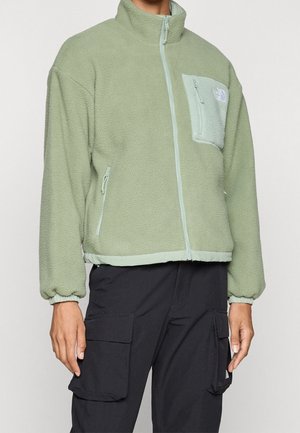 Person wearing light green fleece jacket with front and chest zip pockets, paired with black cargo pants with large side pockets.