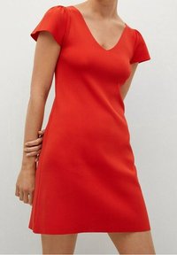 Femme portant une robe rouge vif à manches courtes et décolleté en V, se tenant devant un fond clair, avec une main derrière le dos.