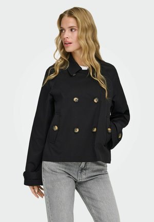 Hosszú szőke hajú nő fekete kétsoros gombos dzsekiben és világosszürke farmernadrágban, oldalra nézve, egyik kezét a zsebében tartva.