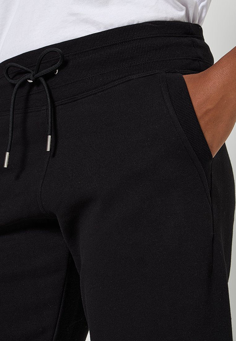 Pantaloni della tuta neri con elastico a coste in vita e lacci, mano nella tasca laterale, abbinati a una maglietta bianca.