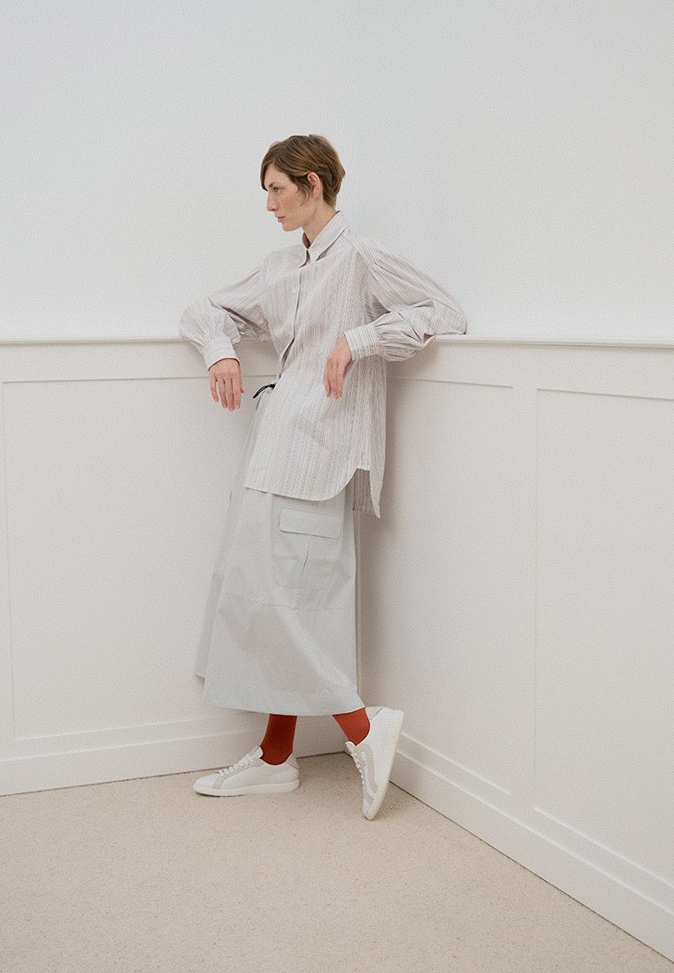 Pasiakowana koszula z guzikami, długa szara spódnica z kieszeniami, białe trampki i czerwone skarpetki. Model opiera się o ścianę. Minimalistyczny design.