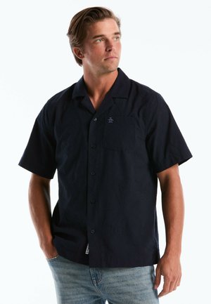 Man met blond haar die een zwart kort mouwen overhemd met knopen draagt en lichtblauwe spijkerbroek, kijkt naar links met één hand in zijn zak.