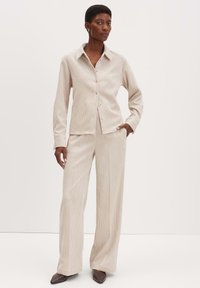 Beige geribbelde button-up shirt gecombineerd met bijpassende wijde broek. De stof heeft een zachte textuur en de outfit heeft een kraag en subtiele knopen.