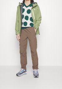Groene waterdichte jas, groen-wit kledingstuk met stippen, bruine broek en grijze sneakers met blauwe accenten. Casual sportoutfit weergegeven.