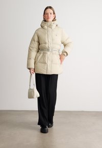 Beige puffjacka med huva, fylld med isolering, med bälte i midjan och logodetaljer. Bärs med svarta byxor och en liten vit väska.