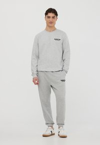Felpa in cotone grigia con joggers abbinati e polsini a costine. Presenta il logo con la scritta "MANHATTAN" in nero sul petto e sulla coscia sinistra.