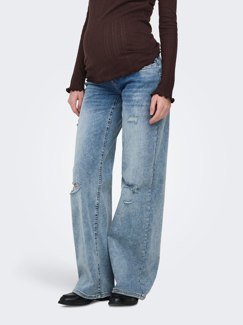 Mannequin portant un jean large déchiré bleu clair, un haut côtelé marron foncé à manches longues et des chaussures noires, debout sur un fond blanc.
