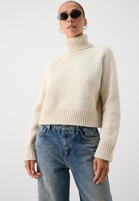 Hvit strikket turtleneck-genser med ribbestrikkede detaljer og en kort design, sammen med blå denimjeans med avslappet passform og falmet effekt.