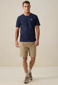Navy katoenen t-shirt met korte mouwen, voorzien van een klein logo, gecombineerd met beige shorts. De outfit is afgemaakt met beige sokken en sneakers.