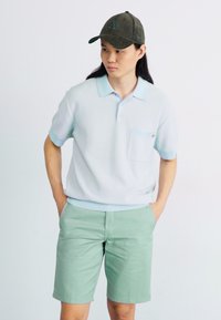 Polo de color azul claro texturizado con un bolsillo, combinado con pantalones cortos de color verde menta. El modelo lleva una gorra de color verde oscuro.