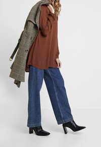 Bruine lange mouwen top gecombineerd met wijde denim jeans en zwarte enkelhoge hakken. Een geruiten overjas hangt over de schouder.