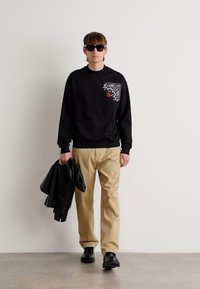 Sudadera negra con un estampado decorativo en el pecho, pantalones beige y zapatos negros. El modelo sostiene una chaqueta negra. Fondo simple.